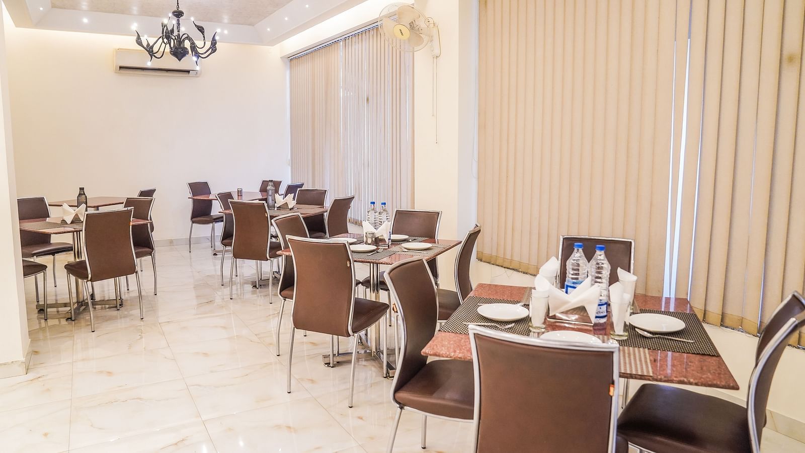 An elegant dining area with multiple tables and chairs is illuminated by a modern chandelier and wall lights - Asian Suites Cyber Park, Gurgaon