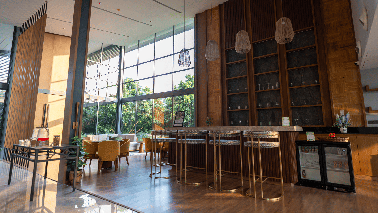 A sophisticated bar area at Wyndham Garden featuring a long counter with high stools, a large built-in wooden shelving unit, and natural light streaming through floor-to-ceiling windows with outdoor views.