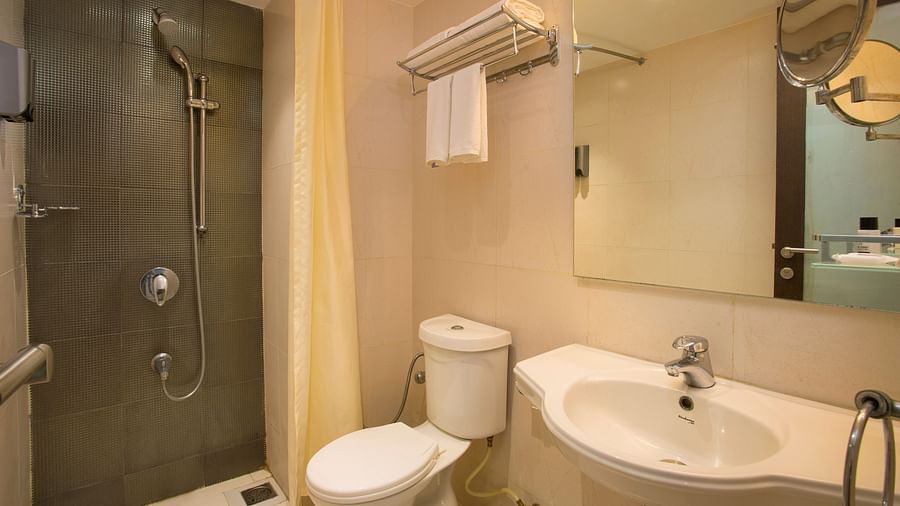 A hotel bathroom with a beige tiled shower area, white toilet, sink and a mirror, with a yellow shower curtain - Hotel Langford Keys by TGI, Shanti Nagar