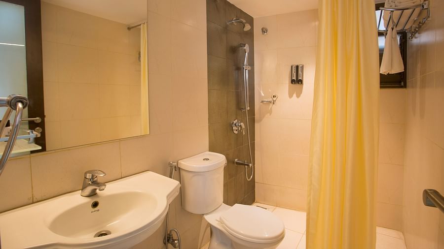 Another view of a hotel bathroom with a beige tiled shower area, white toilet, sink and a mirror, with a yellow shower curtain - Hotel Langford Keys by TGI, Shanti Nagar