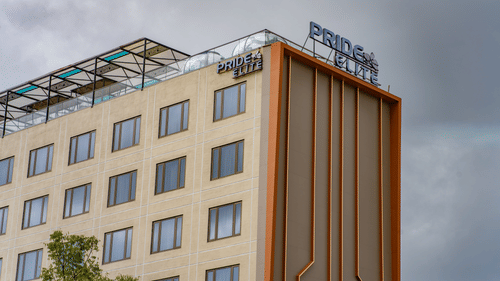A low-angle exterior shot of a modern, multi-storey hotel building with a glass and beige facade, showing the hotel's sign on the roof against an overcast sky