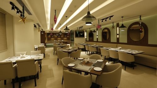  A poster of a restaurant at Tulip Inn, Zirakpur featuring several sets of tables, chairs, and sofa, lights hanging from the ceiling and mirrors attached on the wall