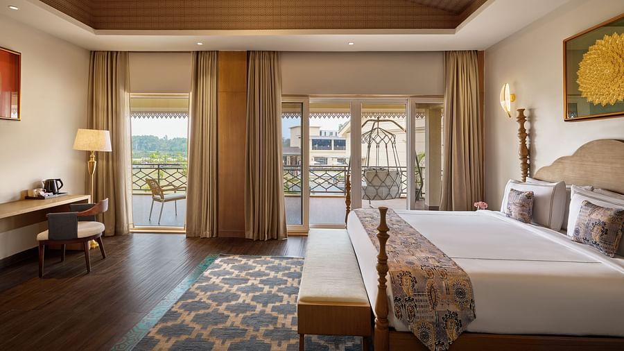 A well-lit hotel room featuring a king size bed with a patterned throw, a wooden desk and chair, and a balcony with a swing offering a view of a cityscape. The room has a wooden ceiling and a patterned rug - Voyage The Solitaire Bliss.