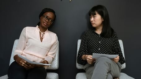 Two women are seated, facing each other in conversation, against a dark background. 