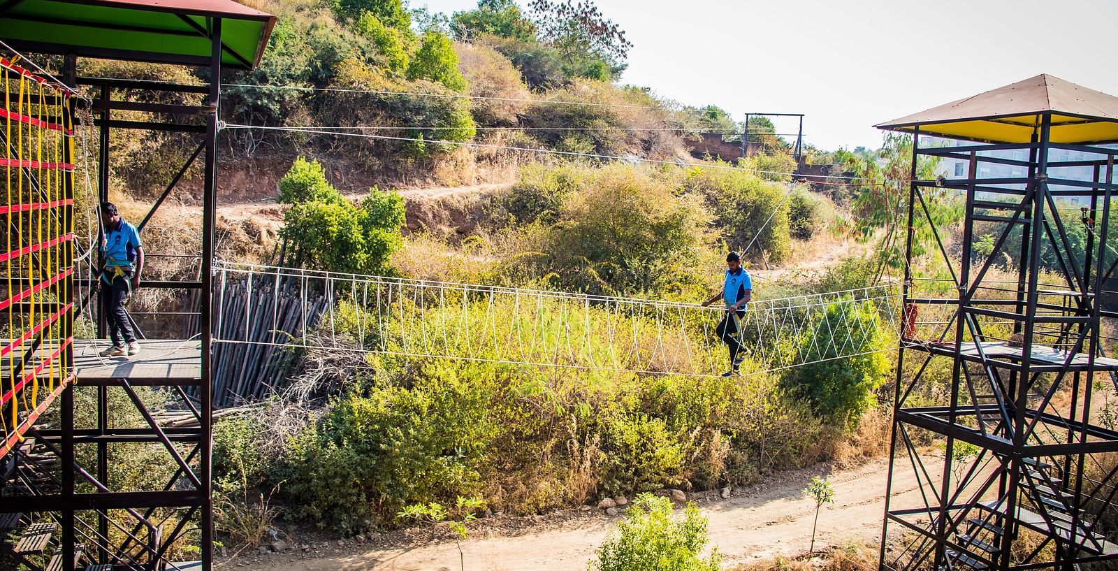 Two people zip-lining across a scenic landscape with hills and trees.- Bamboo Saa Resort & Spa, Udaipur