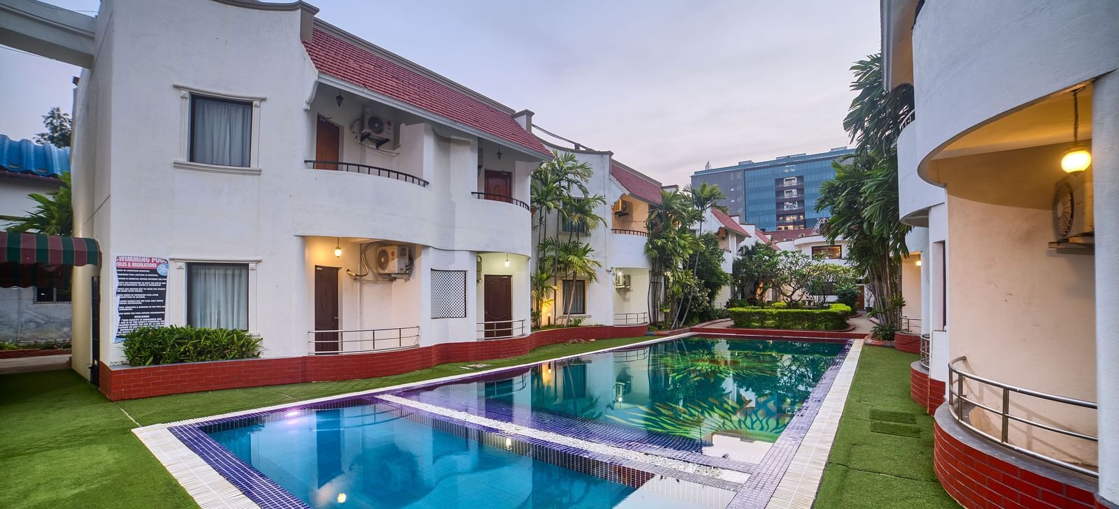 Wide exterior view of the hotel building overlooking a sparkling blue swimming pool surrounded by green lawn at Ilara Hotels & Spa, OMR, Chennai.