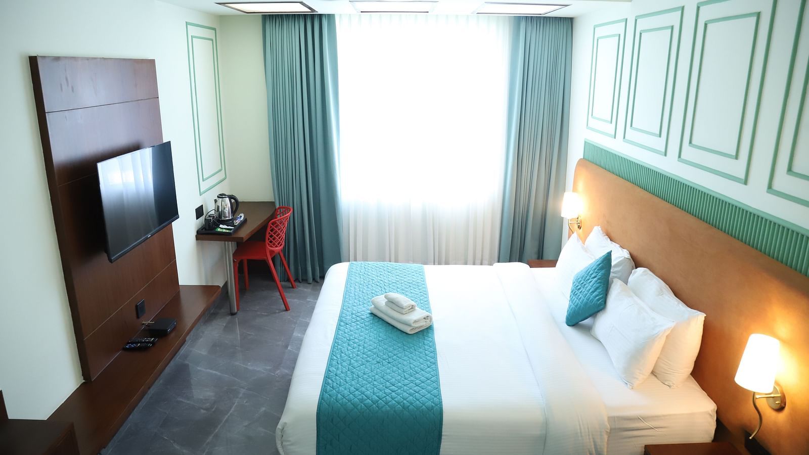 A bright room featuring a double bed with a blue runner, bedside tables, and a television - Asian Suites IFFCO Metro, Gurgaon