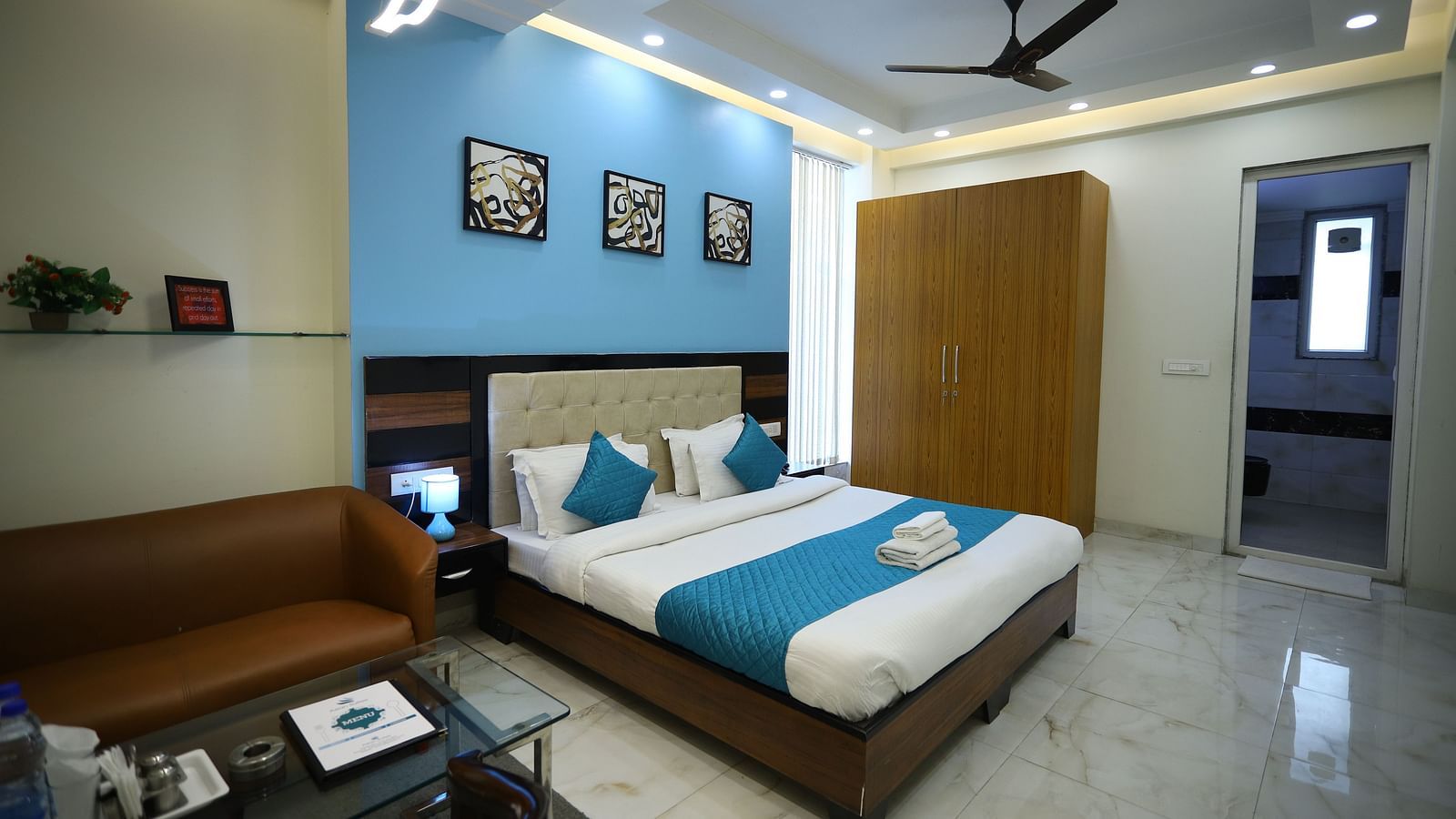 A room with a double bed, a sofa, a coffee table, a ceiling fan, and framed artwork on the wall - Asian Suites Cyber Park, Gurgaon