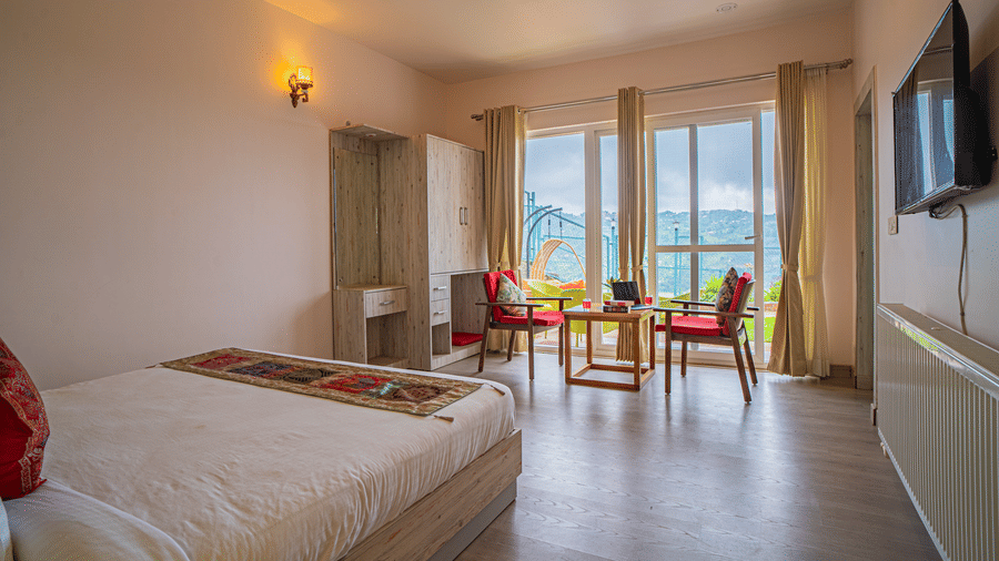 A bedroom with a king size bed, table, chairs, television and a balcony area visible through glass doors at Adrushya Estates, Mukteshwar.