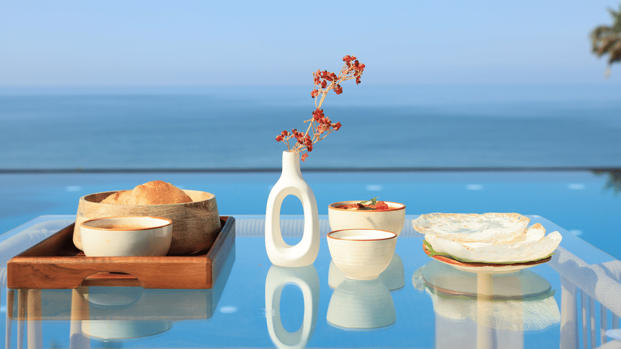 2 bowls of food and a ceramic vase with flowers arranged on a glass table next to an infinity pool overlooking the ocean, at SUKAVI.