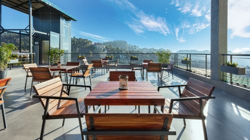 Outdoor dining area at Suryavilas Luxury Resort and Spa, featuring wooden tables and chairs on a spacious terrace with panoramic views of the Himalayan hills under a bright blue sky.