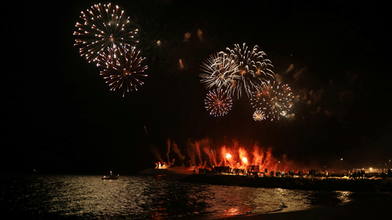 Fireworks explode in the night sky above a body of water reflecting the lights of a nearby dock or shore.