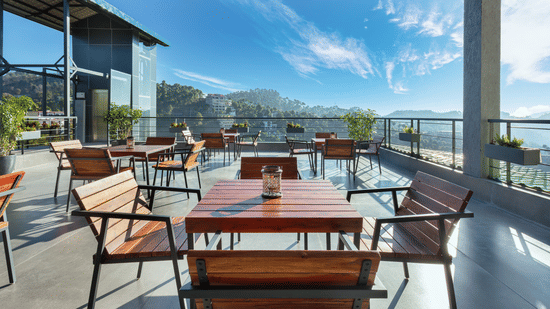Outdoor dining area at Suryavilas Luxury Resort and Spa, featuring wooden tables and chairs on a spacious terrace with panoramic views of the Himalayan hills under a bright blue sky.