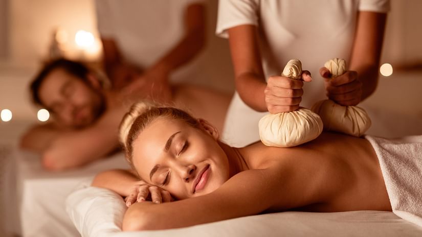 A couple undergoing Ayurvedic kizhi massage, laying on white beds, assisted by two practitioners. 