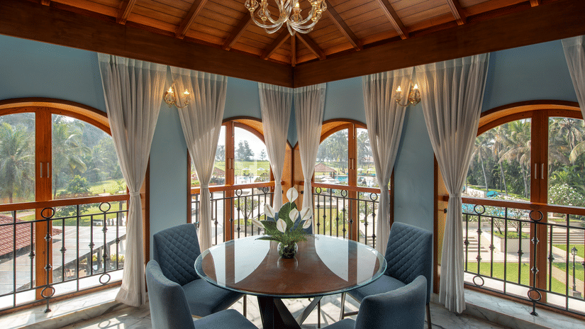 A balcony dining area in the Presidential Suite with a wooden ceiling, light blue walls, a round glass table with a floral centrepiece, and two blue armchairs at Kenilworth Resort & Spa, Goa.