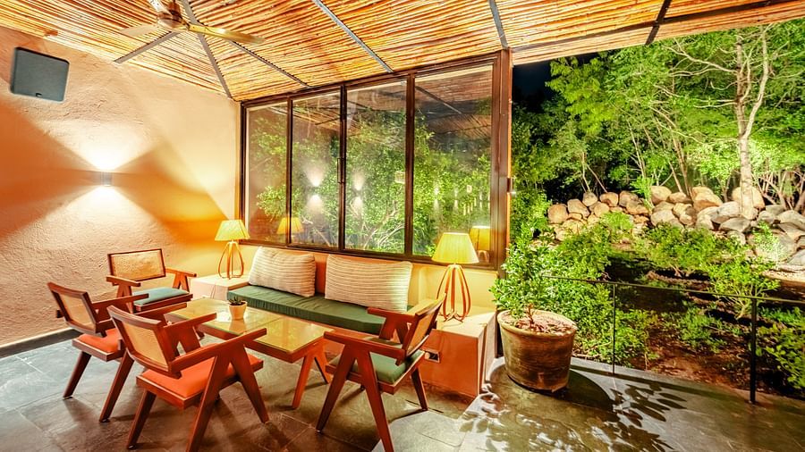 An image showing wooden chairs and table placed aesthetically inside a room with bamboo roofing, where the garden view is at an enjoyable distance