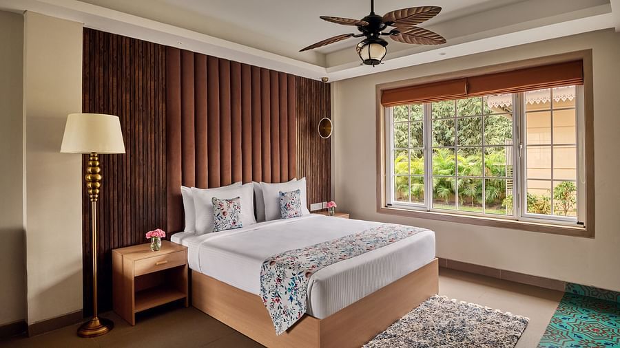 A bedroom with a king bed featuring white linens and a patterned throw, flanked by a wooden nightstand with a lamp and a textured brown headboard. A ceiling fan hangs above, and a large window with blinds reveals a view of lush greenery outside at Voyage T