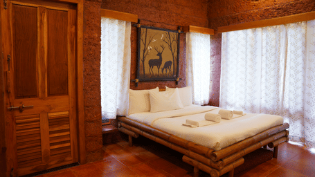 View of a room at Agreen Hotels with a wooden bed frame, rustic wooden interiors, sheer white curtains, and warm natural light.
