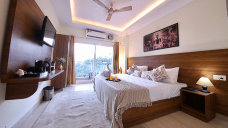 A bedroom at Perfectstayz Value Forest Valley with a king size bed with a white duvet, an A/C, a wall-mounted TV, a nightstand with a lamp, towels in the shape of an animal and sliding glass doors leading to a balcony or view.