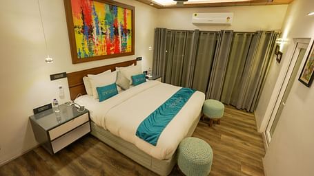 Bedroom of Pent House at The Tattva, Joshimath with a large bed, teal accents, abstract wall art, wooden flooring, grey curtains, and ambient ceiling lighting.