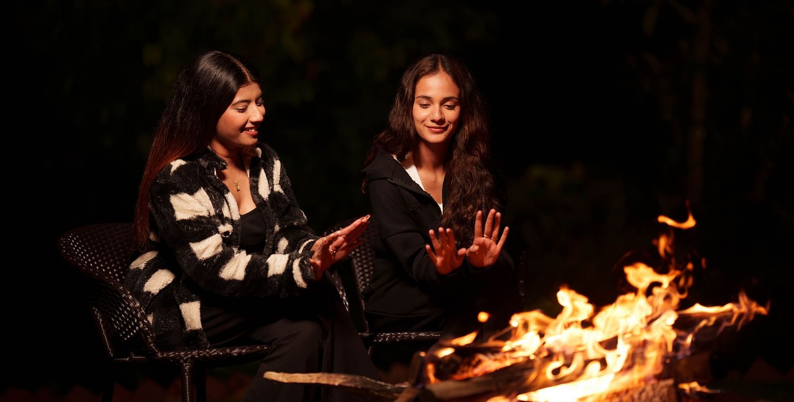 Two women are gathered around a campfire at night, smiling and enjoying the warmth, a common evening activity at Summit Enigma Resort and Spa.