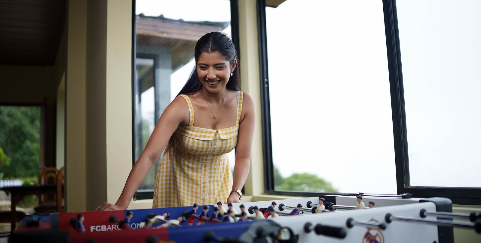 A woman smiling  while playing foosball, with large windows overlooking greenery in the background, possibly in a recreation area at Summit Enigma Resort and Spa.