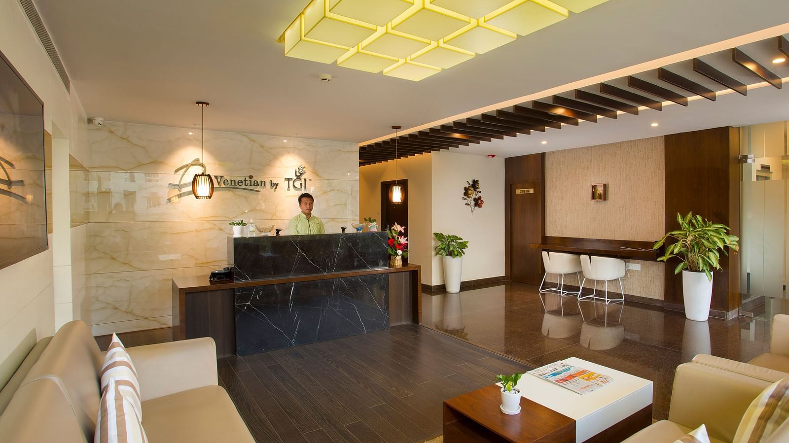 A hotel lobby featuring a marble reception desk, modern seating and geometric ceiling lights - De Venetian by TGI, Brookefield