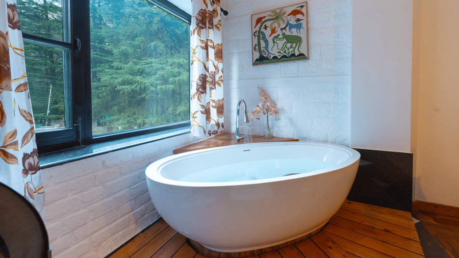 A modern freestanding oval bathtub positioned by a large window with forest views - Johnson Lodge & Spa, Manali
