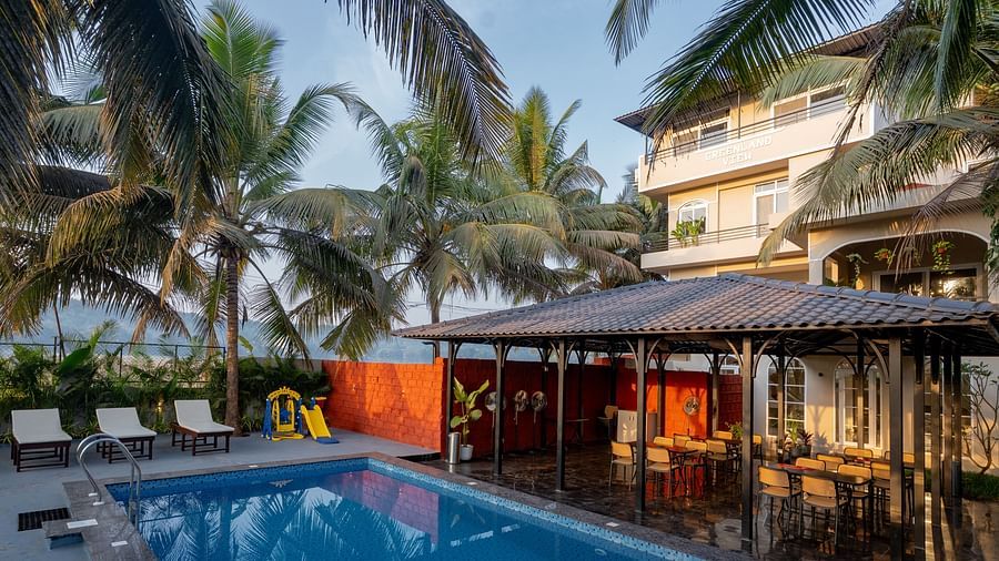Outdoor swimming pool surrounded by palm trees, with a poolside seating area and Kssemya building in the background.