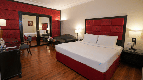 A room at Park Plaza Ludhiana featuring a king bed with white bedding bold red wall panels dark wooden flooring bedside lamps and a seating area near the window.
