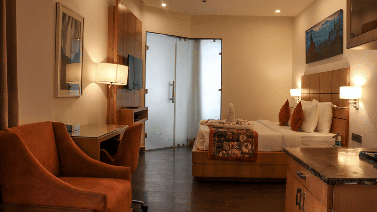 A well-lit, modern hotel room featuring a queen bed with white linens, two orange accent pillows, a decorative throw, and a swan towel art centerpiece.