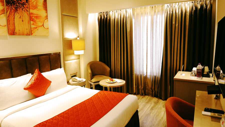 A room at Hotel Saket 27 features a king size bed, upholstered headboard, bedside table, paintings on the wall, desk, chair, television, table lamp and large curtained windows.