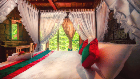 Oak Jacuzzi Villa at Copper Edge - Eco Resort, Coorg, featuring a plush bed with pillows, cushion, and bed runner next to a glass ceiling-to-floor window with greenery view.