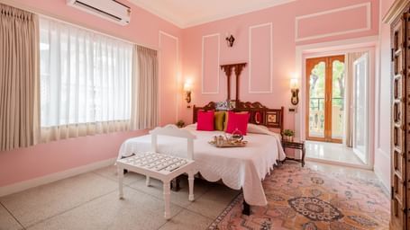 Pink-walled guest room with white bed, decorative cushions, and seating area at Dera Rawatsar, Jaipur.