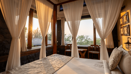 Cozy resort canopy bed view looking out through large glass windows onto a sunset landscape with sitting chairs.