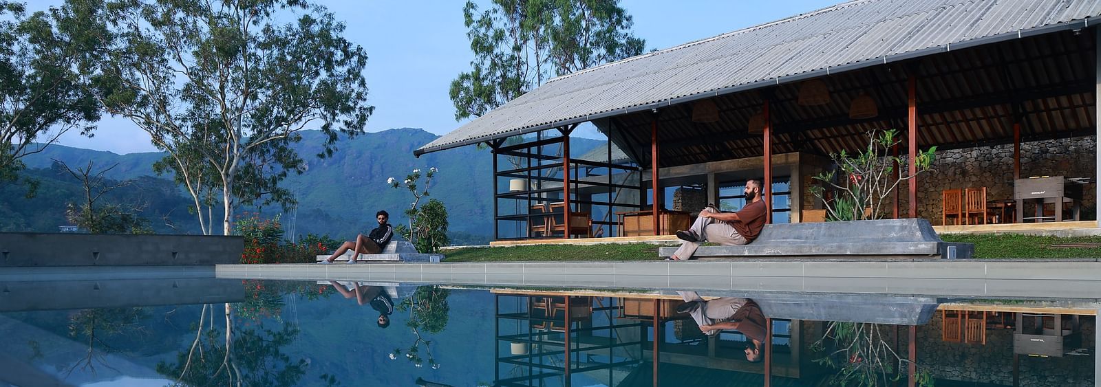 Two people relax by an infinity pool at Vaasta Vagamon, overlooking a serene landscape with rolling hills and trees.