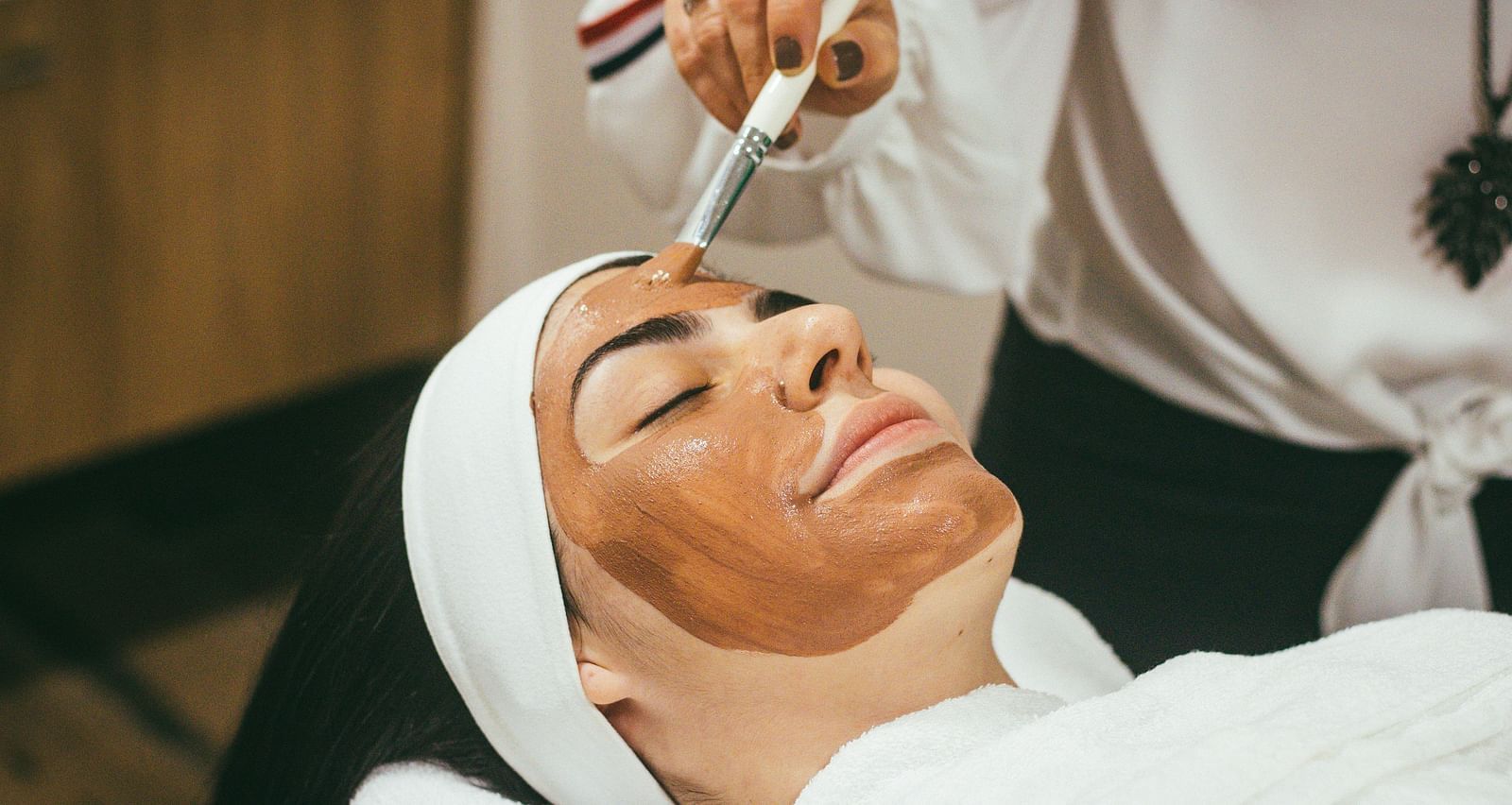 a woman with her eyes closed getting a brown face mask applied on her face
