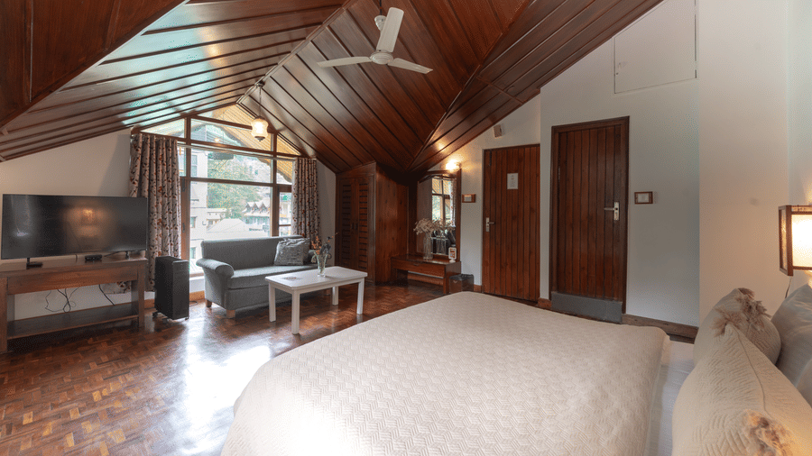 A cosy attic-style room featuring a slanted wooden ceiling, a grey sofa, and a wall-mounted TV - Johnson Lodge & Spa, Manali