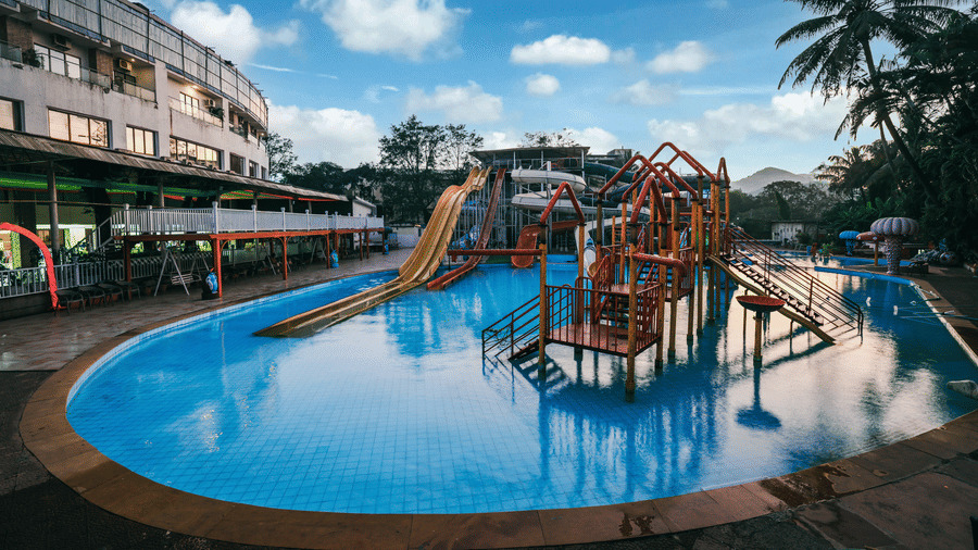 Wide view of a large swimming area and a central water slide island structure surrounded by palm trees and the resort complex at Kumar Resort by Turtle.