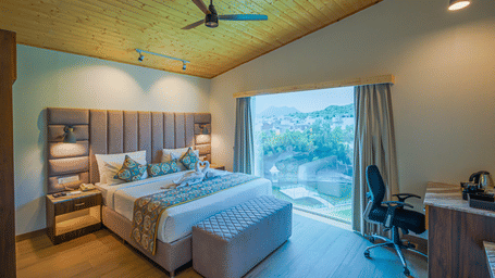 A room at Bamboo Saa Resort & Spa, Udaipur, featuring a large bed, bedspreads, pillows, a fan. a table, and windows with curtains.