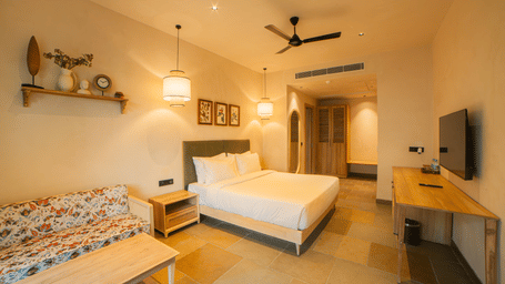 A room with a king size bed, a floral patterned sofa with a light wood coffee table, a wall shelf, and a ceiling fan in a brightly lit space at Jivana Wellness Retreat, Bhopal.