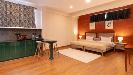 An open-plan studio apartment showing the green cabinetry of the kitchenette and the sleeping area with warm lighting - Johnson Lodge & Spa, Manali