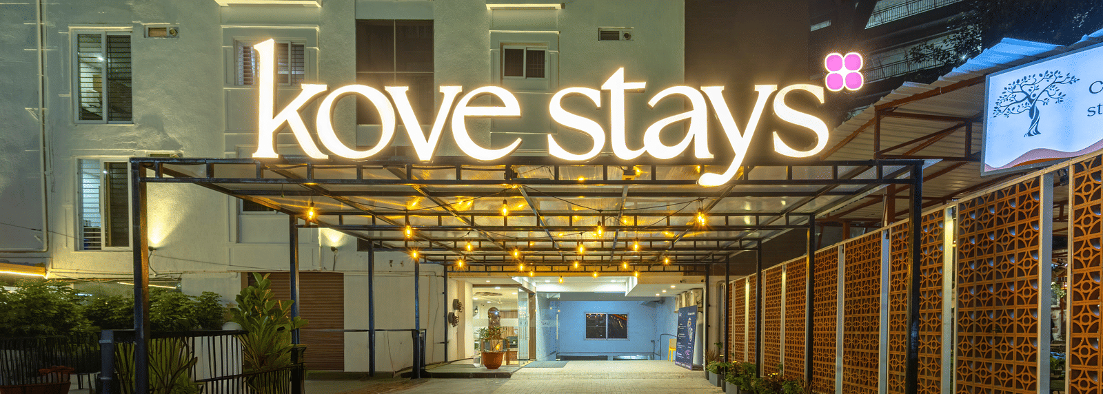 The night view of the building entrance showing a covered walkway with string lights and a large, illuminated 'Kove stays' sign at Kove Smart Stays.