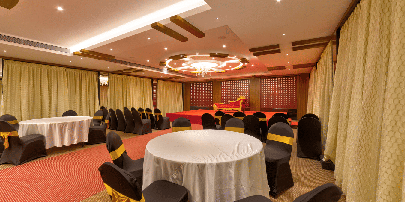 Round tables, black and gold chairs, a red carpet, and a stage setup inside the banquet hall of Icon Express By Bhagini, Hoodi.