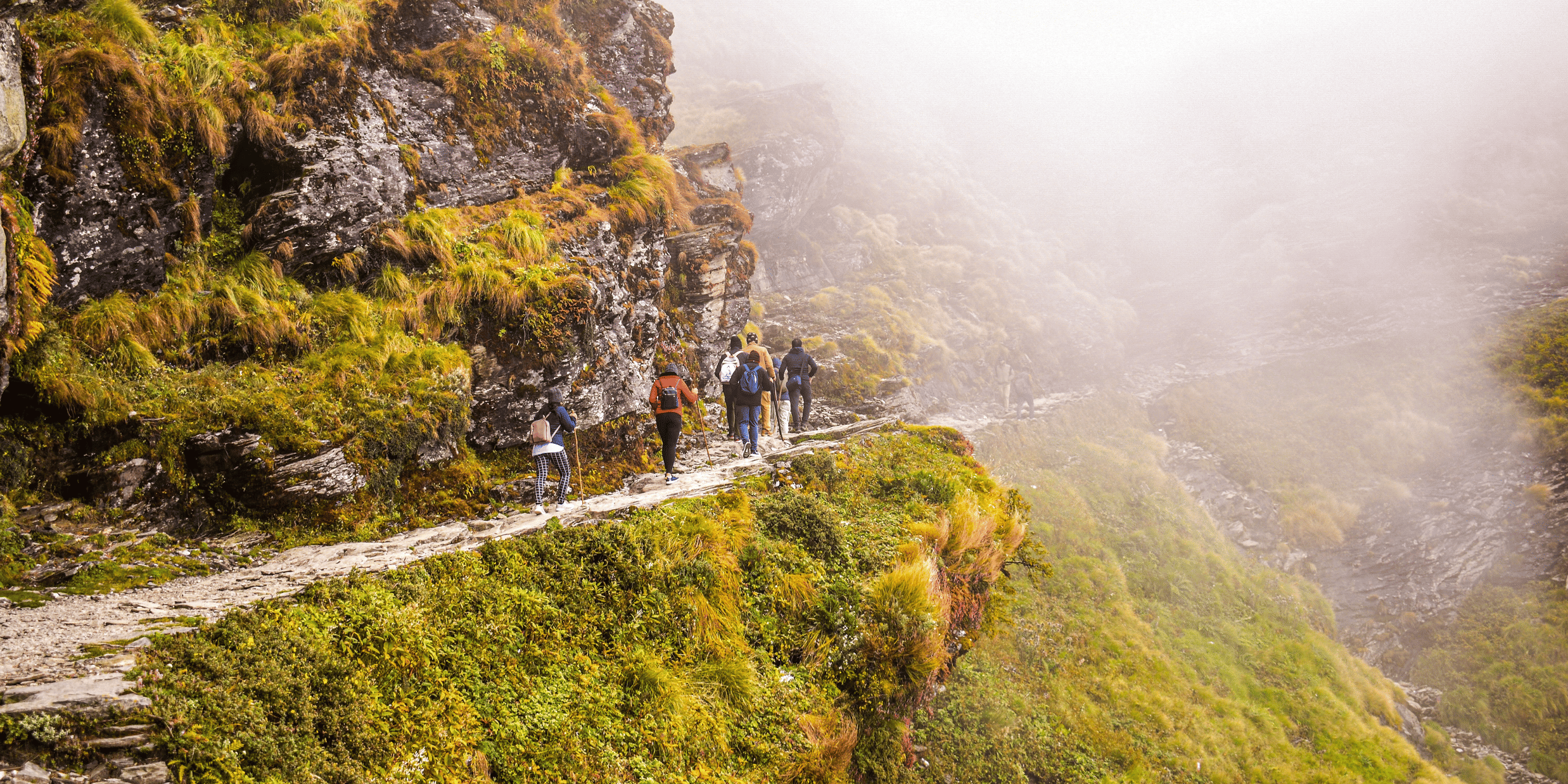 A group of people walking on a rocky hiking trail leading to the misty valleys of a mountain range.