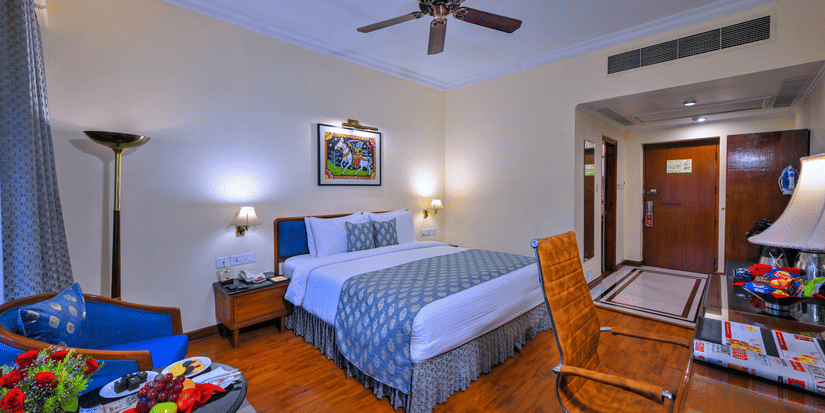 A room with a large bed, spreads, pillows, a fan, and windows at The Clarks Varanasi.