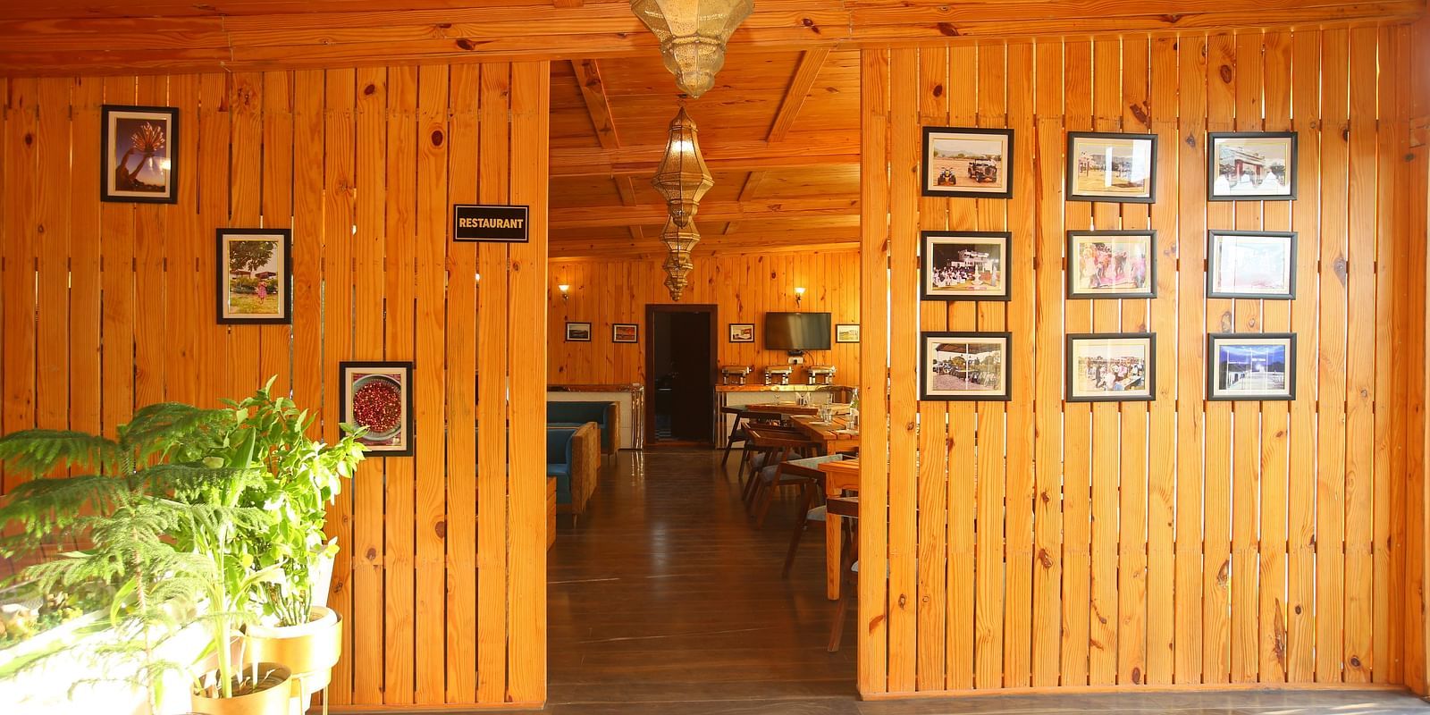 Entryway to the dining area featuring wall-mounted photo frames, a hanging lamp, and potted plants on the side at Aanandam Resort & Spa