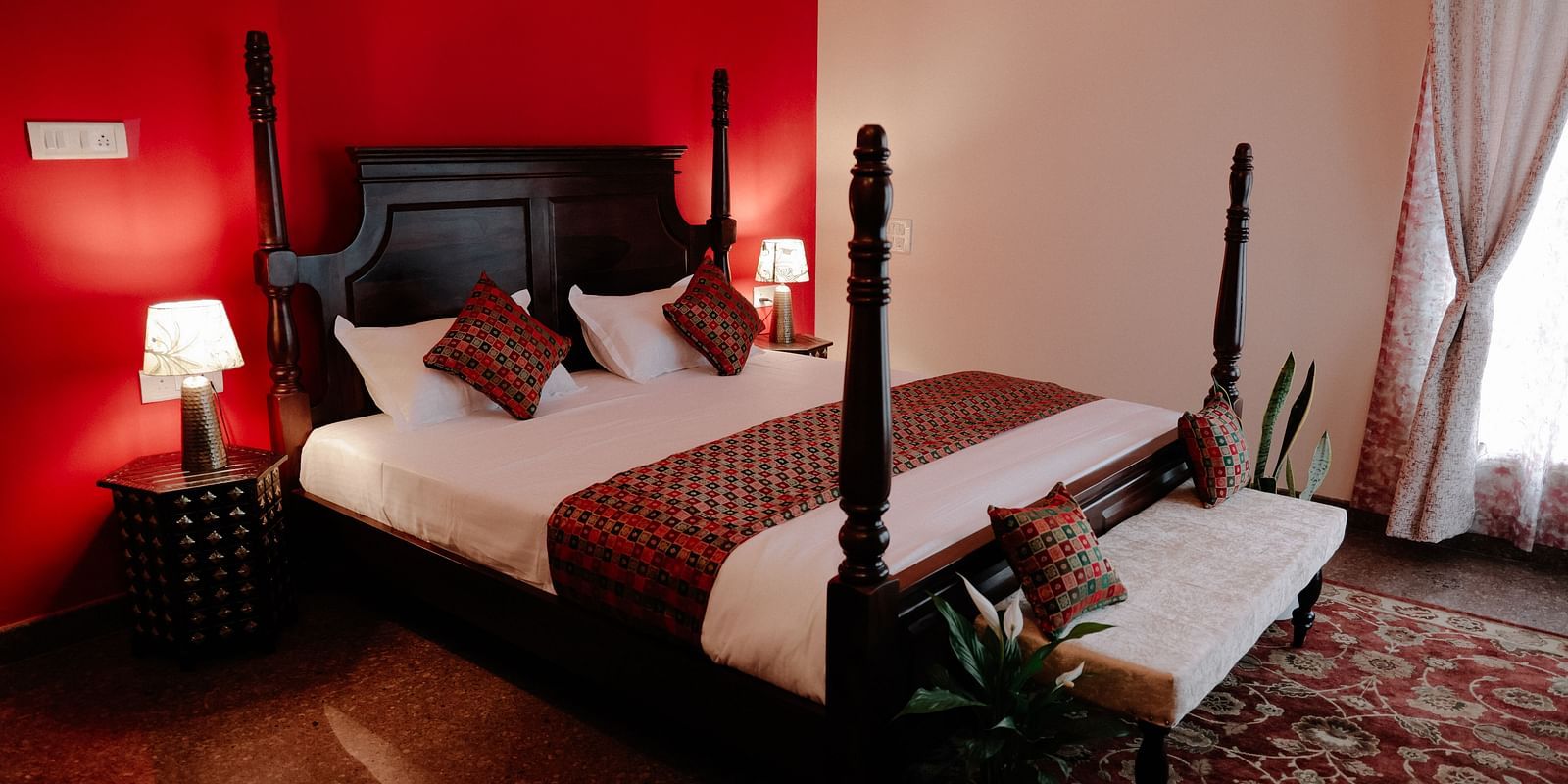 A dark wood four-poster bed with white linens and red patterned throw pillows in a room with a bold red accent wall and a lighter adjacent wall | Adhyaya Heritage Reverbed