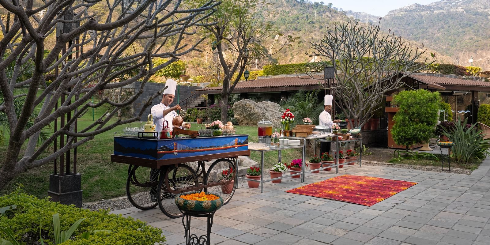 Two chefs are preparing food at outdoor food stations. There are mountains in the background and lush surrounding the scene.