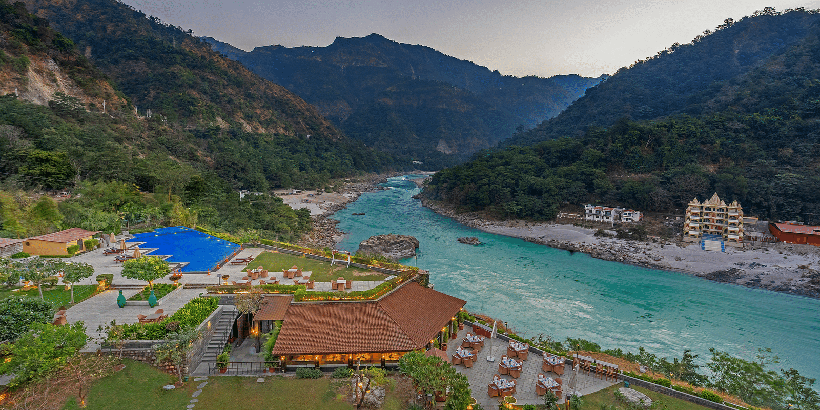 A panoramic view of Aloha on the Ganges, Rishikesh. The image shows the resort nestled amidst hills, with the Ganges River flowing peacefully in the foreground. There is a swimming pool, restaurant, and other facilities visible.